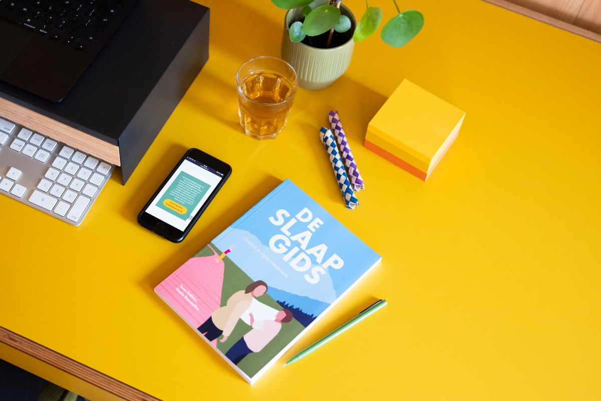 Boek De Slaapgids op bureau met kantoorartikelen.